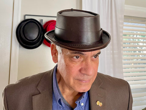Man wearing a dark brown leather pork pie hat with other hats in the background