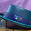 Turquoise leather pork pie hat with a blue hat band and a turquoise jewel against a purple backdrop