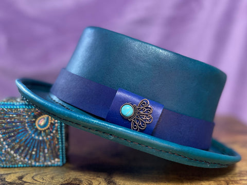 Turquoise leather pork pie hat with a blue hat band and a turquoise jewel against a purple backdrop