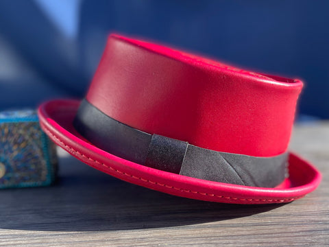 Red leather pork pie hat with a black hat band against a blue backdrop