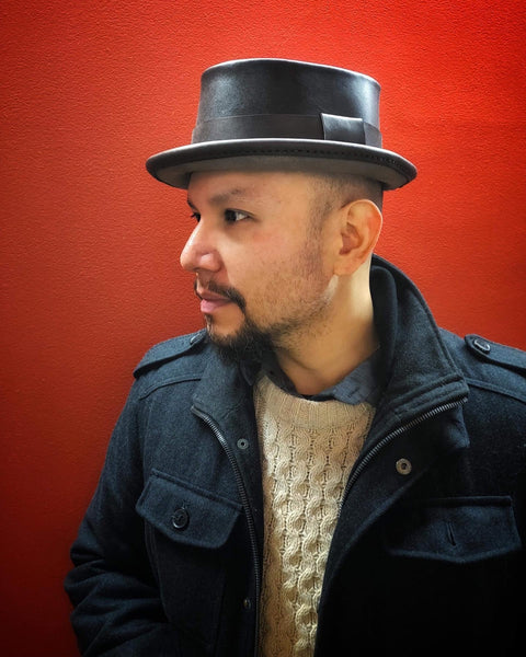 Man wearing a dark brown leather pork pie hat against a red backdrop