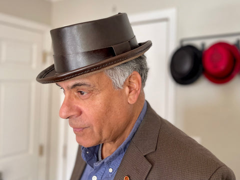 Man wearing a dark brown pork pie hat with other hats in the background