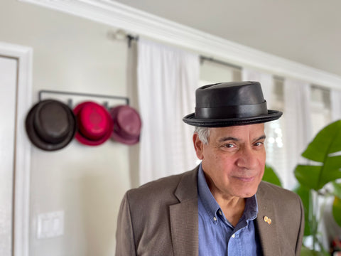 Man wearing a black leather pork pie hat with additional leather pork pie hats on the wall